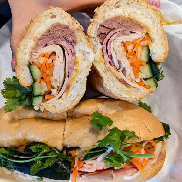 Two Vietnamese banh mi sandwiches, one cut in half, revealing meats, pickled vegetables, and cilantro.