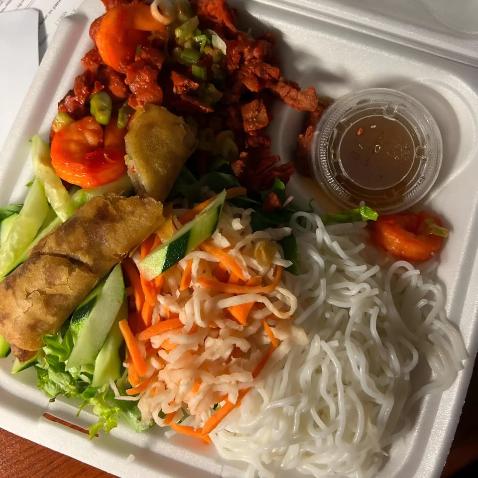 Vietnamese vermicelli bowl with red meat, spring roll, pickled vegetables, and sauce in a takeout container.